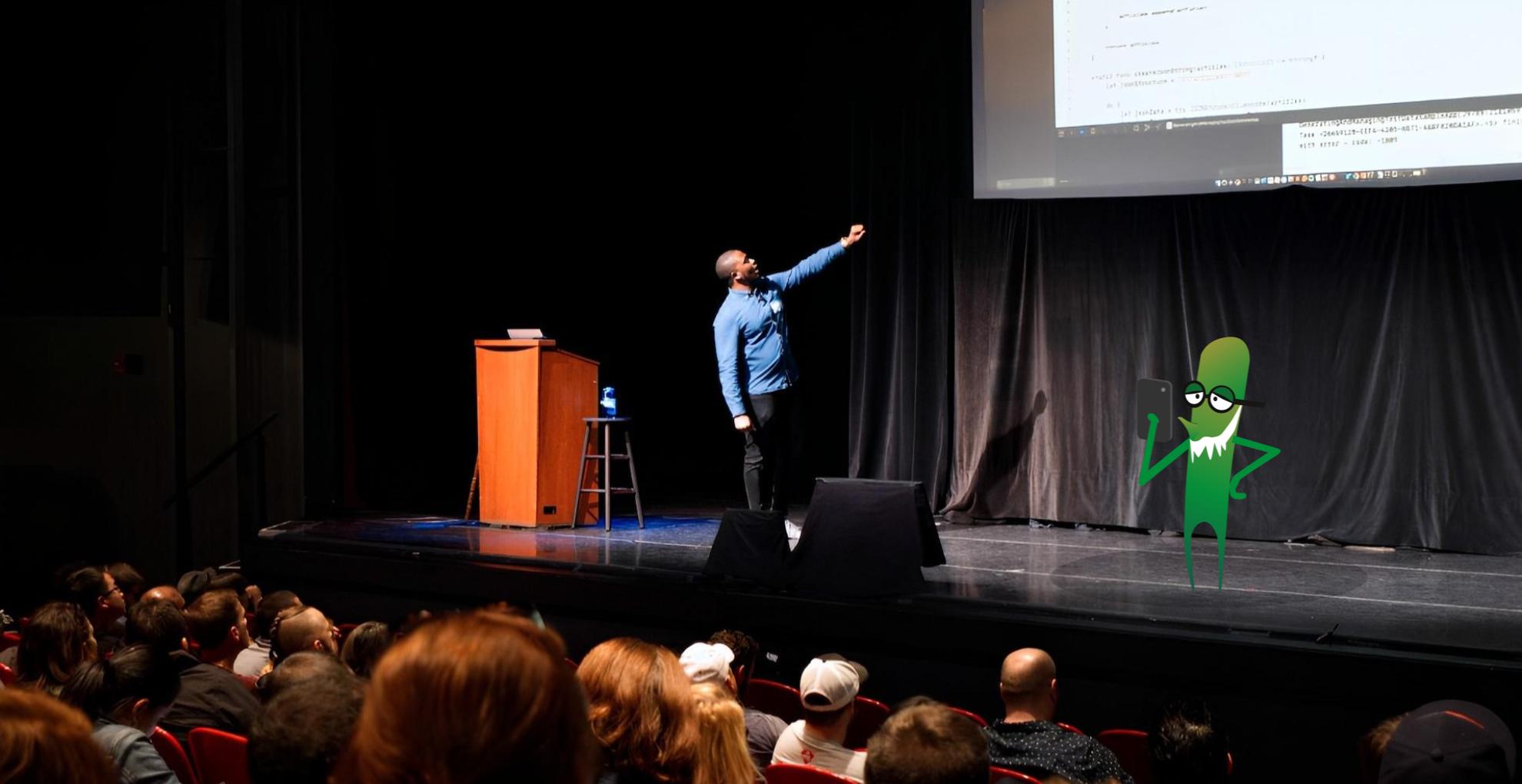 A black man presenting on a big stage. There are people sat in the audience with the back of their heads on show. There is a green monster character on the stage with a mobile phone in its hand.